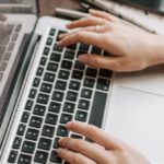 Independent Publishers - From above of unrecognizable woman sitting at table and typing on keyboard of computer during remote work in modern workspace